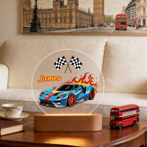 Puede incluir: Luz de noche LED personalizada con un coche deportivo azul, llamas, banderas a cuadros y el nombre "James". La luz est&aacute; sobre una base de madera. Hay un modelo de autob&uacute;s de dos pisos rojo en la mesa.