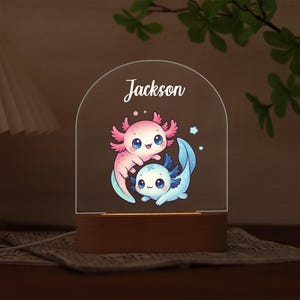 Op de afbeelding: Een decoratief nachtlampje met twee cartoon axolotls, één roze en één blauw, met de naam "Jackson" in script. Het licht heeft een houten voet en een doorzichtige acryl bovenkant.