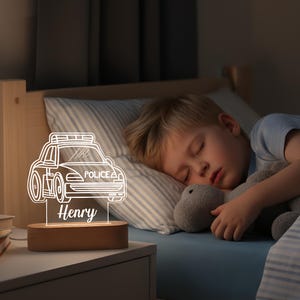 May include: A personalized night light featuring a police car design. The light is made of clear acrylic and sits on a wooden base. The name "Henry" is displayed below the car. The light is on a nightstand next to a bed.