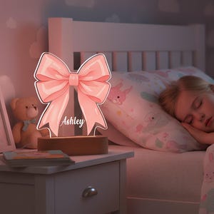 Op de afbeelding: Een roze strikvormig nachtlampje met de naam "Ashley" in script. Het licht staat op een houten voet en verlicht een kinderkamer. Een teddybeer en een boek staan op het nachtkastje.