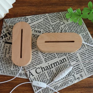 Puede incluir: Dos bases de l&aacute;mpara de madera sin terminar, una ovalada y otra rectangular, con ranuras para luces, descansan sobre una tela con estampado de peri&oacute;dico. Tambi&eacute;n se ve un cable blanco con un interruptor y un panel de control.