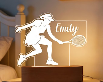 Op de afbeelding: Een tennis-thema nachtlampje met een wit silhouet van een tennisser in actie, met een racket. De naam "Emily" wordt weergegeven in elegant schrift. Het licht staat op een rechthoekige, houten basis en creëert een warme gloed.