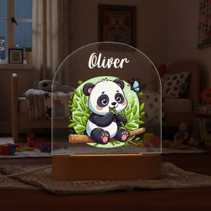 Pode incluir: Uma luz noturna com base de madeira e topo transparente em arco. O topo apresenta um urso panda de desenho animado sentado em um galho, cercado por folhas verdes e uma borboleta azul. O nome "Oliver" está escrito acima do panda.