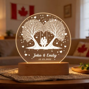 Op de afbeelding: Ronde nachtlamp met een houten voet. Het acryl paneel toont een boomontwerp met een schommel en twee figuren, omringd door harten. De namen "John & Emily" en de datum "12.25.2022" staan eronder gegraveerd.
