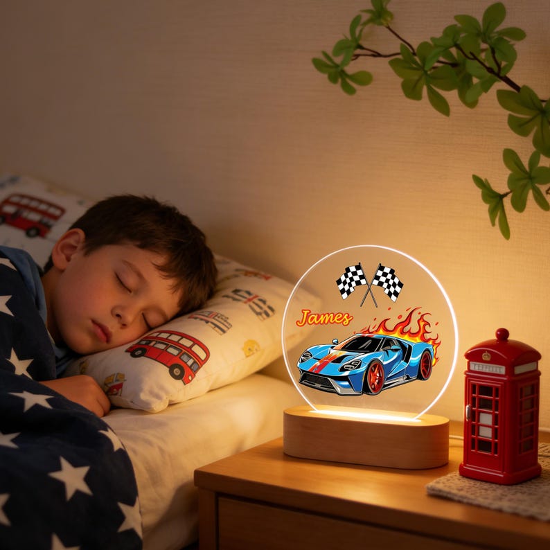 Puede incluir: Una luz de noche con una base de madera y un panel acr&iacute;lico circular transparente. El panel presenta un dise&ntilde;o de coche de carreras azul con llamas, banderas a cuadros y el nombre "James". Una cabina telef&oacute;nica roja est&aacute; a la derecha.