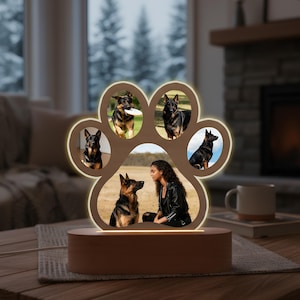 Könnte beinhalten: Ein hölzerner Pfoten-Fotorahmen mit fünf Bildschlitzen, die jeweils einen Hund oder eine Person mit einem Hund zeigen. Der Rahmen wird von einem warmen Schein beleuchtet und steht auf einem Holzsockel.