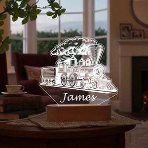 Peut inclure: Lampe de chevet en forme de train blanc avec le nom "James" gravé. La lumière émane d'une base en bois. Le design représente une locomotive à vapeur classique avec des roues et une cheminée détaillées, idéale pour une chambre d'enfant.