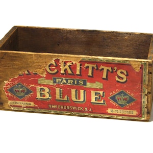 May include: A vintage wooden crate with a faded red and blue label. The label reads "Kitt's Paris Blue" and "New Brunswick, N.J." The crate is rectangular and shows signs of wear, with visible nails and aged wood.