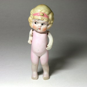 May include: A porcelain doll with blonde hair and a pink dress. The doll has a painted face with blue eyes and a pink headband with a gold accent.