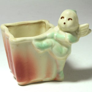 May include: A ceramic planter shaped like a book with a green and white fairy figurine holding the book open. The planter has a pink and white glaze.