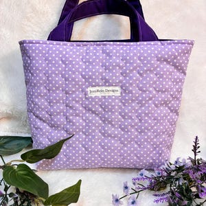 May include: A quilted tote bag in a light purple with white polka dots. The bag has a solid purple handle and trim. The label "JoniRein Designs" is visible on the front of the bag. The bag is sitting on a white, textured surface with greenery and purple flowers.