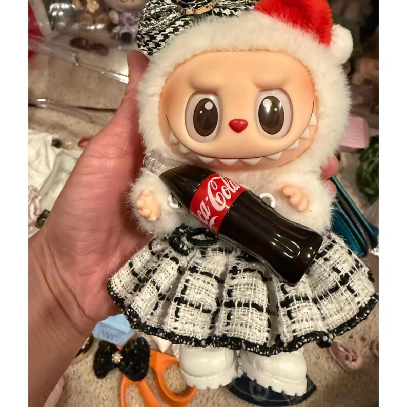 Puede incluir: Una mu&ntilde;eca caprichosa con una capucha de piel blanca con un gorro de Pap&aacute; Noel rojo y una falda estampada en blanco y negro. La mu&ntilde;eca sostiene una botella de Coca-Cola en miniatura. La mu&ntilde;eca tiene ojos grandes y una peque&ntilde;a nariz roja.