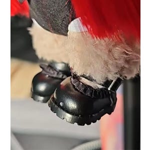 Puede incluir: Primer plano de zapatos de mu&ntilde;eca negros de estilo Mary Jane, con una correa y una suela texturizada. Los zapatos son usados por una mu&ntilde;eca con un atuendo rojo y blanco.