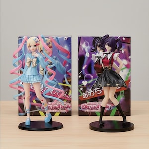 Könnte beinhalten: Zwei Anime-Figuren. Eine trägt ein blaues Outfit mit Schleife, die andere ein rotes und schwarzes Kleid. Beide stehen auf schwarzen Sockeln vor Kartons mit japanischem Text und farbenfrohen Grafiken. Die Figuren stehen auf einer hellbraunen Oberfläche.