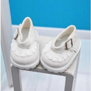 Puede incluir: Un par de zapatos blancos de mu&ntilde;eca estilo Mary Jane. Los zapatos tienen un ribete con volantes alrededor de la puntera y un peque&ntilde;o cierre de hebilla. Est&aacute;n sobre una superficie blanca con un fondo azul.