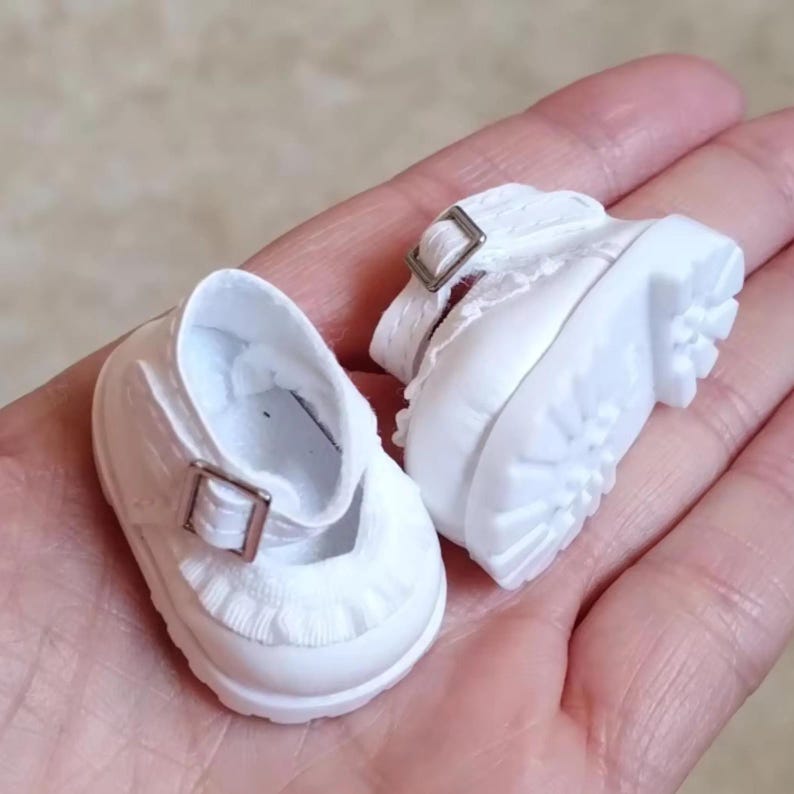 Puede incluir: Un par de zapatos blancos de mu&ntilde;eca estilo Mary Jane. Los zapatos tienen un cierre de hebilla y un ribete con volantes alrededor de la abertura. Las suelas son gruesas y texturizadas.