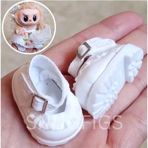 Puede incluir: Un par de zapatos blancos de mu&ntilde;eca estilo Mary Jane, con una correa y hebilla plateada. Los zapatos tienen una suela texturizada y un ribete con volantes. Una peque&ntilde;a mu&ntilde;eca con un vestido blanco est&aacute; en el fondo.