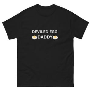 Peut inclure: T-shirt noir avec l'inscription blanche "DEVILED EGG DADDY" et deux illustrations d'œufs mimosa. Le t-shirt est de style col rond.
