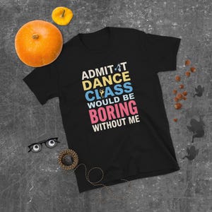 May include: Black t-shirt with the text "ADMIT DANCE CLASS WOULD BE BORING WITHOUT ME" in white, yellow, blue, and pink. The shirt is displayed on a gray surface with a pumpkin, small gourd, glasses, and a decorative rope.
