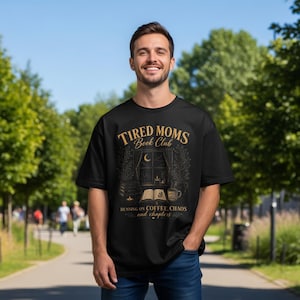 Peut inclure: T-shirt noir avec l'inscription dorée "Tired Moms Book Club". Le motif comprend une scène de fenêtre avec une lune, un livre et une tasse de café. Le texte "Running on coffee, chaos and chapters" est en dessous, pour les amateurs de lecture.