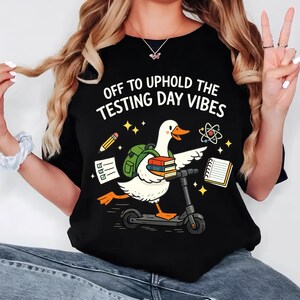Peut inclure: T-shirt noir avec l'inscription "OFF TO UPHOLD THE TESTING DAY VIBES". Le motif représente une oie blanche sur un scooter, portant des livres et un sac à dos. D'autres graphiques incluent un crayon, des étoiles et un symbole d'atome.
