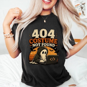 Puede incluir: Una camiseta negra con un gráfico de un fantasma con sudadera con capucha escribiendo en un portátil. El texto dice 404 COSTUME NOT FOUND. El diseño incluye un fondo de atardecer naranja con siluetas de murciélagos volando y estrellas en el cielo.