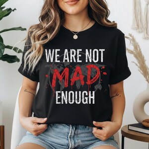 Peut inclure: T-shirt noir avec le texte blanc "WE ARE NOT" au-dessus du texte rouge "MAD" et du texte blanc "ENOUGH". Le mot "MAD" a un design vieilli et fissuré. Un motif de carte du monde est derrière le texte.
