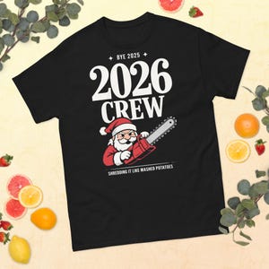 Peut inclure: T-shirt noir avec le texte "BYE 2025" et "2026 CREW". Un dessin animé du Père Noël tient une tronçonneuse. Le texte "Shredding it like mashed potatoes" est en dessous. Le t-shirt est entouré de fruits.