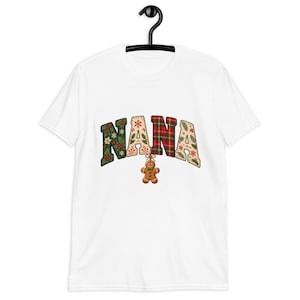 May include: A white short-sleeved t-shirt featuring the word NANA in large, arched block letters. Each letter has a unique Christmas-themed pattern including holly, snowflakes, and red plaid. A small gingerbread man hangs from the letter N.
