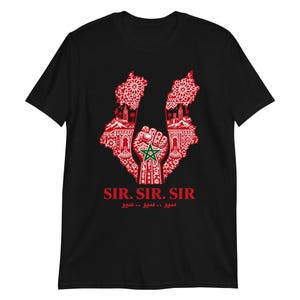 Peut inclure: Un t-shirt noir à manches courtes avec un motif rouge représentant un poing levé au centre d'une carte du Maroc. Le design inclut des motifs complexes, des silhouettes architecturales et le drapeau marocain. Le texte SIR. SIR. SIR. est inscrit.