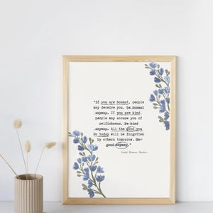 May include: A framed print with a light wood frame. The print features a quote in black text with blue floral accents. A beige vase with dried flowers sits to the left of the frame. The quote reads, "If you are honest, people may deceive you. Be honest anyway."