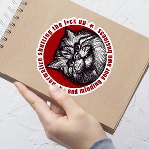 Puede incluir: Una pegatina con una ilustración en blanco y negro de la cara de un gato dentro de un círculo rojo. El texto alrededor del círculo dice: "normaliza callarte y ocuparte de tus asuntos."