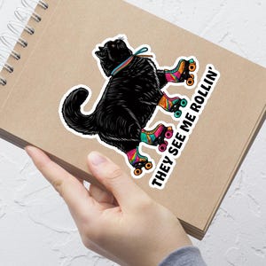 Puede incluir: Un gato negro con patines de ruedas de colores. El gato mira hacia la derecha y el texto "They see me rollin'" está escrito en la pegatina.