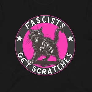 May include: Black t-shirt featuring a graphic of a black cat with claws out, inside a pink circle with the text "Fascists Get Scratches". The design includes stars and is printed on a short-sleeved tee.