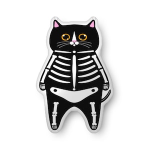 May include: A black cat-shaped pillow with a white skeleton design. The cat has yellow eyes, pink ears, and a pink nose. The skeleton design includes ribs, spine, and leg bones. The pillow is a Halloween-themed decorative item.