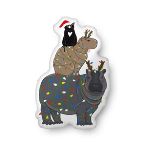 May include: A whimsical Christmas-themed pillow featuring a black cat wearing a Santa hat and a heart on its chest, perched atop a capybara and a hippo, both adorned with festive string lights and reindeer antlers. The pillow is white.
