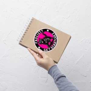 May include: A sticker featuring a black cat with the text "Fascists Get Scratches" in a circular design. The cat is in a dynamic pose, set against a pink background with white stars. The sticker is on a brown spiral notebook.