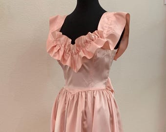 May include: A vintage-style, pale pink dress with a ruffled neckline and a full skirt. The dress is displayed on a black mannequin. The fabric appears to be a smooth satin or similar material, with a slight sheen. The dress has a fitted bodice and a flared skirt.