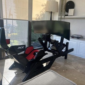 May include: A black and red racing simulator setup with a curved monitor. The seat is black with red accents, and the frame is black. The text "RCC" is visible on the side of the seat. The setup is on a tiled floor.