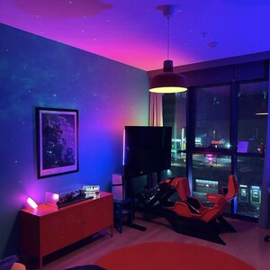 May include: A modern gaming room with a red and black racing simulator chair, a large flat-screen TV, and a red cabinet. The room has a starry sky effect on the ceiling and walls, with a red circular rug on the floor. A framed picture hangs on the wall.