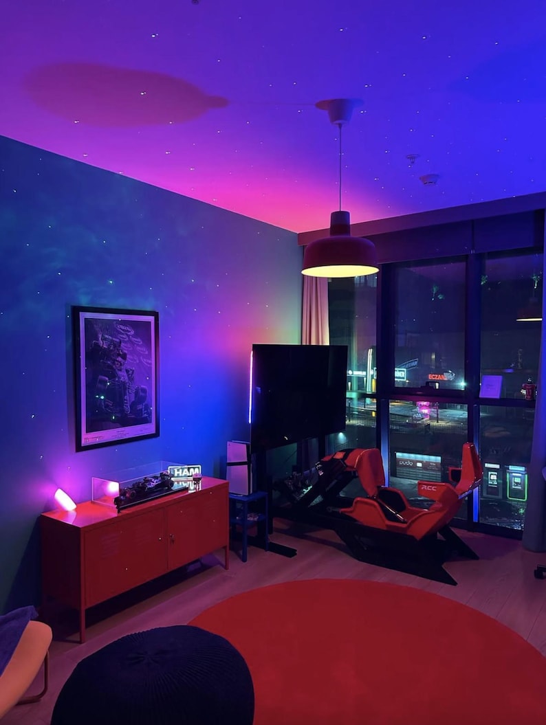 May include: A modern gaming room with a red and black racing simulator chair, a large flat-screen TV, and a red cabinet. The room has a starry sky effect on the ceiling and walls, with a red circular rug on the floor. A framed picture hangs on the wall.