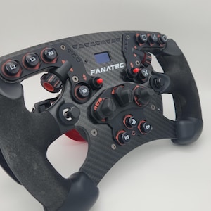 Op de afbeelding: Een zwart en rood race stuurwiel met een carbonfiber afwerking. Het stuurwiel heeft meerdere knoppen en een klein LCD-scherm waarop "FANATEC" wordt weergegeven.