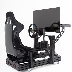 May include: Black racing simulator with a black racing seat, a steering wheel, a shifter, and a monitor. The simulator is on wheels and has a black metal frame. The simulator is labeled "Dyno ARC".