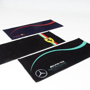 May include: Three rectangular black towels with Formula 1 team logos. One towel has a Red Bull Racing logo, another has a Ferrari logo, and the third has an AMG Petronas Formula One Team logo with a Mercedes-Benz emblem.