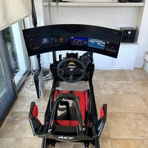 May include: A black and red racing simulator setup with a curved monitor displaying a racing game. The simulator features a steering wheel, pedals, and a racing seat. A gaming console is visible to the left. The setup is designed for immersive racing experiences.