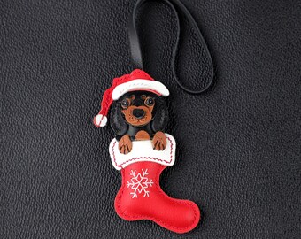 May include: A leather Christmas ornament depicting a dachshund wearing a Santa hat, emerging from a red stocking. The stocking has a white cuff and a snowflake design. A black strap is attached for hanging.