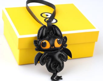 May include: Black leather dragon keychain with orange eyes and small wings. The dragon is attached to a black leather loop. The keychain is in front of a yellow box with a white lid.