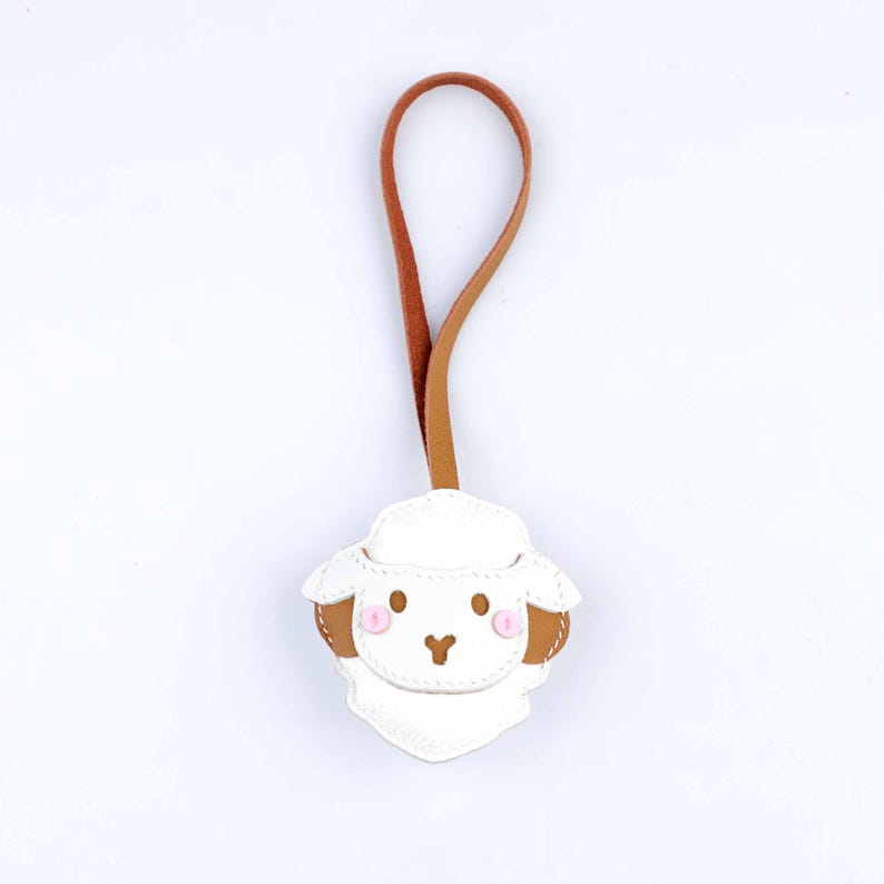 May include: A white leather sheep-shaped keychain with brown ears and a pink blush. The keychain has a brown and tan leather loop for hanging. The sheep has a stitched design and a cute, cartoonish appearance.
