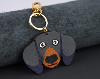 May include: A dachshund-shaped keyring with a gold-coloured metal clasp and ring. The dog's head is black, with a brown snout and white-rimmed eyes. The ears are a darker blue colour.