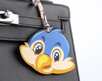 May include: A blue, yellow, and tan bird-shaped leather bag charm. The charm is attached to a black leather handbag with a patterned fabric strap. The bird has large, expressive eyes and a detailed beak.